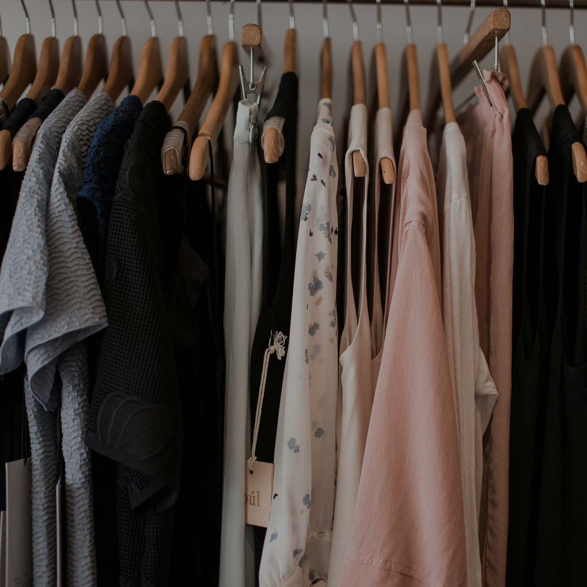 A close-up view of a clothing rack holding multiple garments on wooden hangers, including gray, black, cream, and blush pink tops and dresses.