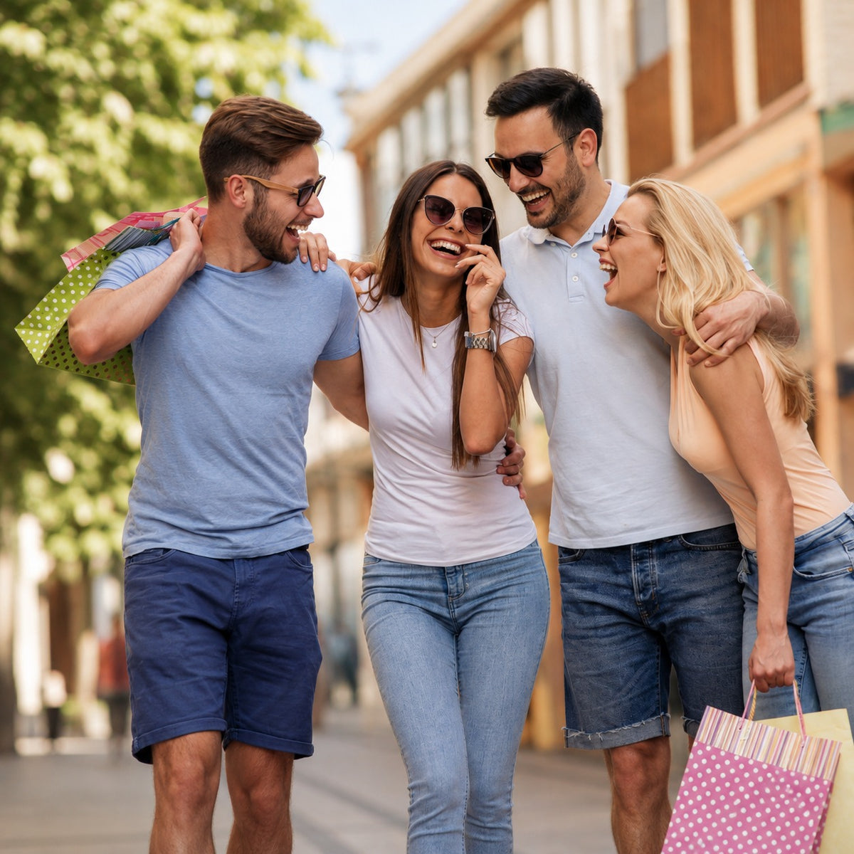 Four happy young adults walking and laughing on a sunny city street, each carrying a pink shopping bag. They are dressed in casual summer clothing, with two wearing blue jeans and two wearing denim shorts.