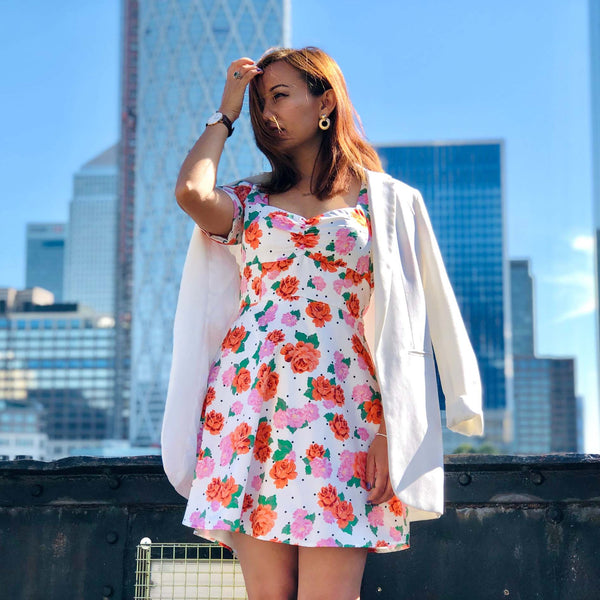 A woman stands outdoors in front of a city skyline wearing a white sleeveless dress with a red, orange, and pink floral print, styled with a white blazer draped off her shoulders.