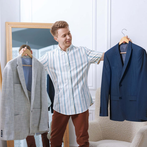 A man stands indoors holding three suits on hangers: a light gray blazer on the left, a navy blue blazer on the right, and a light blue striped button-down shirt in the center. A mirror and upholstered chair are visible in the room.