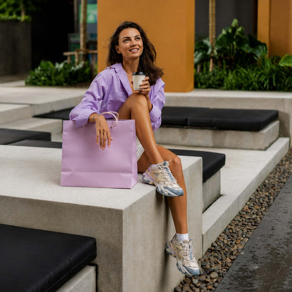 A woman sits on outdoor concrete steps holding a light purple shopping bag and a takeaway coffee cup. She is wearing a lavender button-up shirt, white shorts, and metallic silver sneakers.